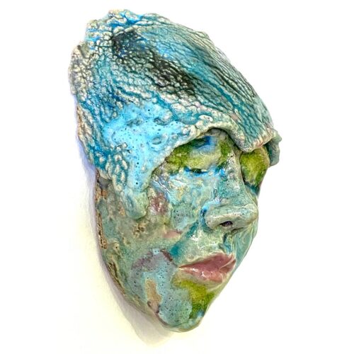 A wall hanging ceramic sculpture of a resting female face. She is covered with a multitude of green, turquoise and pink textured glazes.